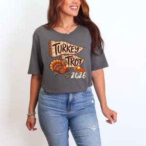 May include: A dark gray short-sleeve t-shirt featuring a graphic of a running turkey wearing sneakers and a headband. Above the turkey are two pennant flags with a tan checkered pattern and the text "TURKEY TROT" and "2026" with a slice of pie.