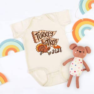 May include: A cream-colored short-sleeve baby bodysuit featuring a graphic of a running turkey wearing a headband and sneakers, two pennant flags with the text TURKEY TROT, a slice of pumpkin pie, and the year 2026 in brown and orange tones.