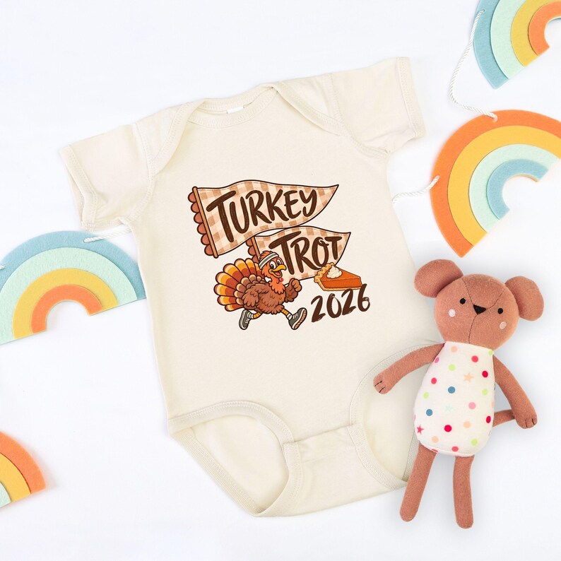 May include: A cream-colored short-sleeve baby bodysuit featuring a graphic of a running turkey wearing a headband and sneakers, two pennant flags with the text TURKEY TROT, a slice of pumpkin pie, and the year 2026 in brown and orange tones.