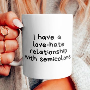Könnte beinhalten: Eine weiße Keramiktasse mit schwarzem Text, der "Ich habe eine Hassliebe zu Semikolons." lautet.
