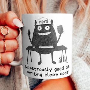 Op de afbeelding: Een witte keramische mok met een zwart-witte illustratie van een monster dat een hoed draagt met de tekst "nerd". Het monster zit aan een bureau met een computer en een telefoon. De tekst onder het monster zegt "Monstrously good at writing clean code!"