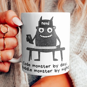 Op de afbeelding: Een witte keramische mok met een zwart-witte illustratie van een monster dat een hoed draagt met de tekst "nerd". Het monster zit aan een bureau met een computer en de tekst "Code monster by day, cuddle monster by night."