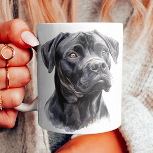 Könnte beinhalten: Weiße Keramiktasse mit einem Aquarellporträt eines schwarzen Hundes mit braunen Augen. Das Fell des Hundes ist mit Schattierungen detailliert. Die Tasse wird von einer Person gehalten, die Ringe und ein Armband trägt.