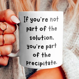 Könnte beinhalten: Eine weiße Keramiktasse mit schwarzem Text, der lautet: "Wenn du nicht Teil der Lösung bist, bist du Teil des Niederschlags."