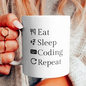 Peut inclure: Une tasse en céramique blanche avec du texte noir qui dit "Eat Sleep Code Repeat". La tasse est tenue par une personne portant un cardigan blanc.