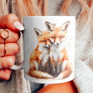 Peut inclure: Une tasse en céramique blanche avec une illustration aquarelle de deux renards en train de se câliner. Les renards sont orange et blanc avec des accents bruns. L'arrière-plan est brun clair avec des mouchetures blanches.