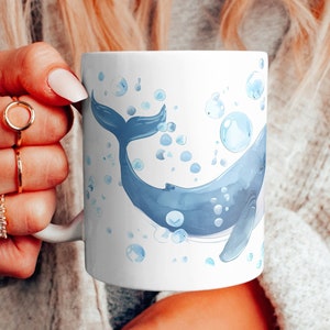 May include: A white ceramic mug with a blue watercolor whale design and blue bubbles. The whale is swimming upwards with its tail in the air.