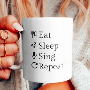 Op de afbeelding: Witte keramische mok met zwarte tekst die "Eten, Slapen, Zingen, Herhalen" luidt.