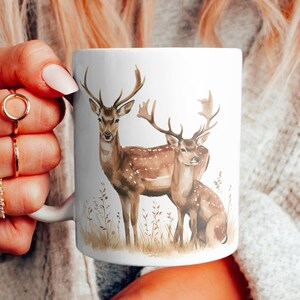 Könnte beinhalten: Weiße Keramiktasse mit einer Aquarell-Illustration von zwei Rehen in einer Waldlandschaft. Die Rehe sind braun mit weißen Flecken und stehen auf einer Wiese mit grünem Gras und braunen Blättern.