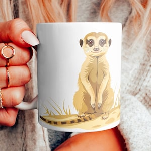 Könnte beinhalten: Weißer Keramikbecher mit einer Illustration eines Erdmännchens. Das Erdmännchen ist in Braun- und Beigetönen dargestellt und sitzt aufrecht. Der Becher hat ein schlichtes, sauberes Design, geeignet für Kaffee oder Tee.