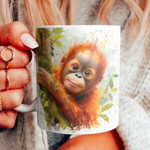 Op de afbeelding: Witte keramische mok met een aquarel-illustratie van een orang-oetan-baby die uit groene bladeren gluurt. De orang-oetan heeft een lichtbruine vacht en een nieuwsgierige uitdrukking.