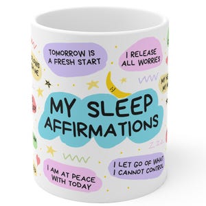 Könnte beinhalten: Eine weiße Keramiktasse mit einem farbenfrohen Design, das den Text "Meine Schlaf-Affirmationen" und positive Affirmationen wie "Morgen ist ein Neuanfang" und "Ich lasse alle Sorgen los" zeigt.
