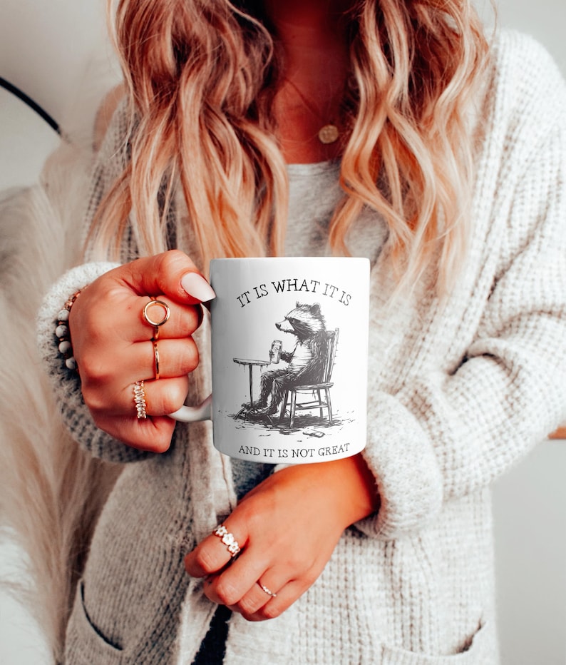 May include: White ceramic mug with a black and white illustration of a raccoon sitting in a chair and holding a glass. The text on the mug reads "It is what it is and it is not great."