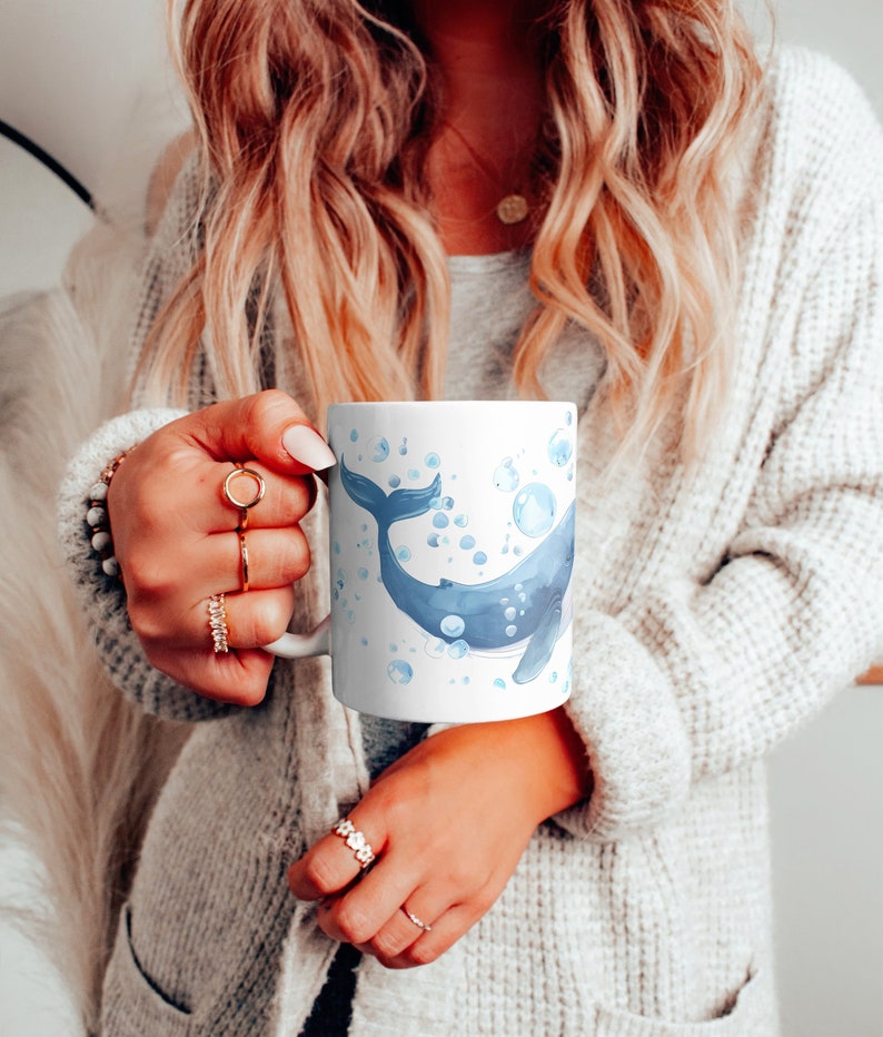 May include: A white ceramic mug with a blue watercolor whale design and blue bubbles. The whale is swimming upwards with its tail in the air.