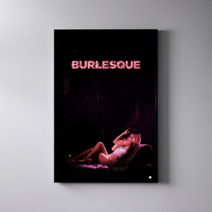 Puede incluir: Una mujer con un traje de burlesque blanco y rosa yace en un escenario con un sombrero negro en la cabeza. El escenario está cubierto de purpurina y la palabra "BURLESQUE" está escrita en luces rosas sobre ella.