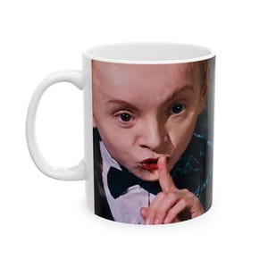 May include: A white ceramic mug featuring a photographic print of a person with a pale complexion, wearing a white shirt and black bow tie, holding a finger to their lips in a shushing gesture. The person has dark eyes and red lips.