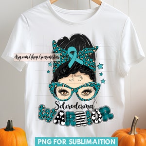 May include: White t-shirt featuring a graphic of a woman with a teal ribbon, glasses, and a leopard print headband. The text "Scleroderma WARRIOR" is below the image. The shirt is hanging on a wooden hanger.