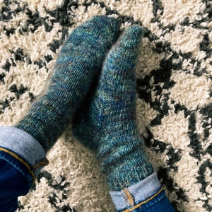 May include: A pair of hand-knitted socks in shades of teal, blue, and green. The socks are worn with rolled-up denim jeans. The socks are resting on a patterned, off-white and gray rug.
