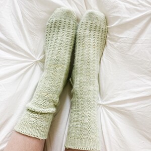 May include: A pair of light green knitted socks with a ribbed texture. The socks are on a white bedspread.