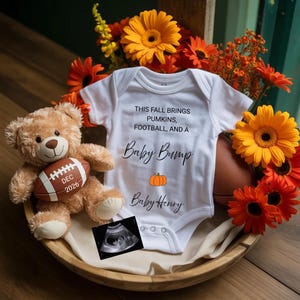 May include: A white baby onesie with the text "THIS FALL BRINGS PUMKINS, FOOTBALL, AND A Baby Bump Baby Henry" and a pumpkin graphic. A teddy bear holding a football with "DEC 2026" is next to the onesie, along with an ultrasound image and orange flowers.