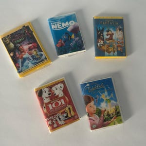 May include: Five miniature VHS tapes featuring Disney animated films. Titles include "The Princess and the Frog," "Finding Nemo," "Fantasia," "101 Dalmatians," and "Tinker Bell: Secret of the Wings." The tapes have colorful covers and yellow plastic cases.