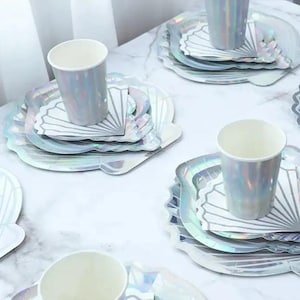 Peut inclure: Ensemble de plats et de gobelets en papier blanc irisé avec un motif de coquillage. Les assiettes sont empilées et les gobelets sont à côté. Les assiettes et les gobelets sont sur une surface en marbre blanc.