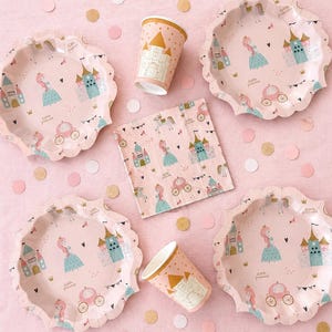 May include: Pink princess-themed party supplies, including scalloped plates, cups, and a napkin. The plates and cups feature a princess, castle, and carriage design. Gold and pink confetti dots the background.