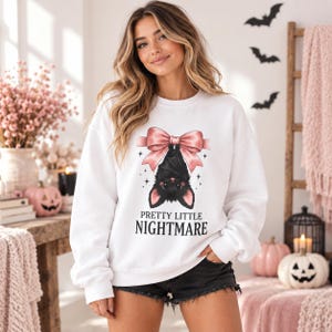Peut inclure: Un sweat-shirt blanc à manches longues avec un motif représentant une chauve-souris noire suspendue à un grand nœud rose. Le texte "PRETTY LITTLE NIGHTMARE" est imprimé en lettres capitales noires sous l'illustration.