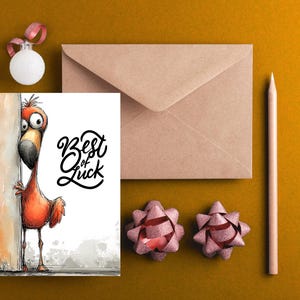 May include: A greeting card with a cartoon flamingo peeking from behind a wall, with the words "Best of Luck" in black script. The card is next to a brown envelope, a pencil, two pink bows, and a white ornament with a pink ribbon.