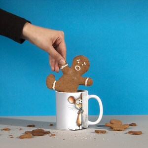 May include: A gingerbread man cookie is held above a white mug with a mouse illustration. The gingerbread man has white icing accents and a surprised expression. Cookie crumbs are scattered on the surface, with a blue background.