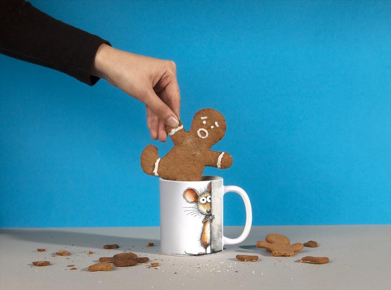 May include: A gingerbread man cookie is held above a white mug with a mouse illustration. The gingerbread man has white icing accents and a surprised expression. Cookie crumbs are scattered on the surface, with a blue background.