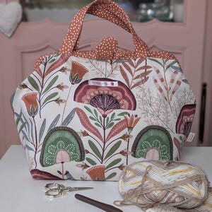 Può includere: Una borsa tote con stampa floreale e manico a pois marroni. La borsa presenta uno sfondo color crema con un design botanico nei toni del verde, marrone e bordeaux. Un gomitolo di lana, forbici e un uncinetto sono davanti alla borsa.