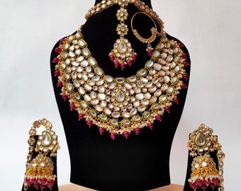 May include: A traditional Indian bridal jewellery set featuring a gold-toned necklace with white and green stones, a matching maang tikka, earrings, and a nose ring. The set is decorated with red beads.
