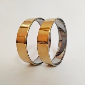 Peut inclure: Deux bracelets dorés à bandes plates. La surface extérieure de chaque bracelet est dorée et brillante, tandis que la bande intérieure est d'une couleur métallique plus foncée. Les bracelets sont présentés sur un fond blanc uni.