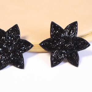 Könnte beinhalten: Ein Paar schwarze Perlenohrringe in Blumenform. Die Ohrringe haben die Form eines fünfzackigen Sterns und bestehen aus kleinen schwarzen Perlen.