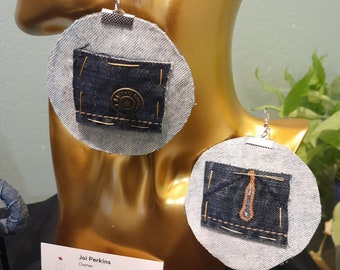 Op de afbeelding: Een paar denim oorbellen met een ronde vorm en een klein vierkantje van denim stof in het midden. De oorbellen zijn gemaakt van een lichtgrijze stof met een subtiele textuur. De denim stof is donkerblauw en heeft een knoop in het midden. De oorbellen hangen aan zilveren haken.