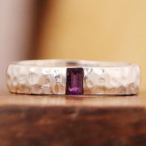 以下が含まれることがあります： ハンマーで叩いたような質感のシルバーのバンドリングに、小さな長方形の紫色のアメジストの宝石が埋め込まれています。