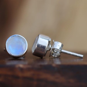 May include: Close-up of two silver stud earrings with round, iridescent moonstone cabochons. The earrings are set on a dark wooden surface, with a blurred background. The moonstones have a soft, milky appearance with a subtle play of color.