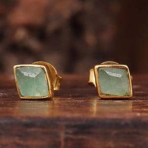 Puede incluir: Un par de pendientes de botón cuadrados. Cada pendiente presenta una piedra preciosa verde claro engastada en un marco de metal dorado. Los pendientes se exhiben sobre una superficie de madera oscura, con un fondo borroso.