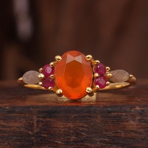 May include: A gold ring with an oval orange gemstone at its centre, complemented by smaller red and grey gemstones. The ring is displayed on a dark wooden surface, highlighting the jewellery's design and colour contrast.