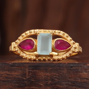 Op de afbeelding: Een gouden ring met een rechthoekige, lichtblauwe edelsteen geflankeerd door twee druppelvormige, rode edelstenen. De ring heeft een textuur, kralenontwerp en een warme, gouden tint. De ring wordt gepresenteerd op een donker houten oppervlak.