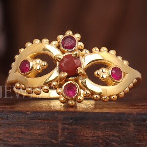 Puede incluir: Un anillo dorado con una gema roja central y gemas rojas más pequeñas. El anillo tiene un diseño detallado con un motivo floral y está sobre un fondo de madera oscura. El texto "LAJARA JEW" es visible.