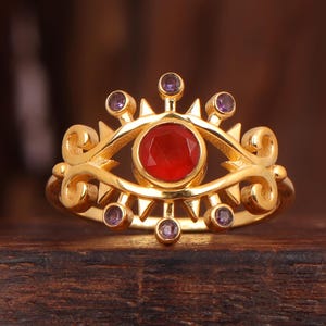 May include: A gold ring with an eye motif, featuring a central red gemstone and smaller purple stones. The ring's design includes decorative swirls. The background is a dark wooden surface.
