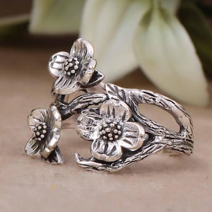Puede incluir: Un anillo de plata con un diseño floral. El anillo presenta tres flores con pétalos detallados y un tallo texturizado.