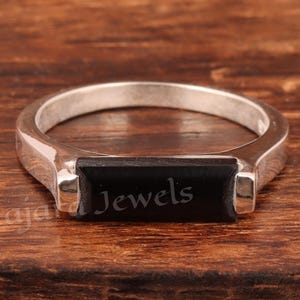 Könnte beinhalten: Ein silberner Ring mit einem rechteckigen schwarzen Stein. Auf dem Ring steht das Wort "Jewels". Der Ring liegt auf einer dunklen Holzoberfläche.