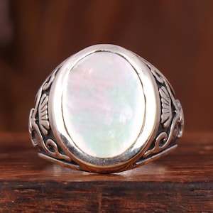 Puede incluir: Un anillo de plata con una piedra ovalada de nácar iridiscente. El anillo tiene un diseño detallado y grabado alrededor de la banda. El anillo está sobre un fondo de madera oscura.