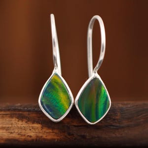 May include: A pair of silver drop earrings with teardrop-shaped, iridescent green and yellow gemstone insets. The earrings are displayed on a dark wooden surface, with a blurred brown background.
