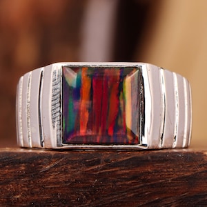 Op de afbeelding: Een zilveren ring met een rechthoekige, veelkleurige edelsteen. De edelsteen toont tinten van rood, oranje, groen en blauw. De ring heeft een geribbeld ontwerp aan de zijkanten, wat een textuurelement aan de sieraden toevoegt.
