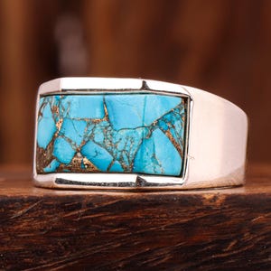 以下が含まれることがあります： 長方形のターコイズの象嵌が施されたシルバーリング。ターコイズは金色の静脈を持つ、割れたような模様です。リングは暗い木製の表面に置かれ、ジュエリーのデザインを際立たせています。