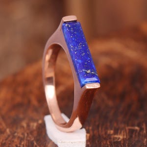 May include: A rose gold ring featuring a rectangular, deep blue lapis lazuli stone. The ring's band has a modern, minimalist design. The lapis stone has flecks of gold, adding contrast to the rich blue color. The ring is displayed on a small white surface.